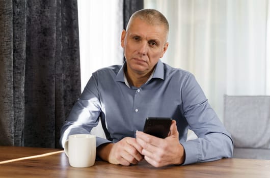 Portrait of a male businessman who sits at a table, holds a phone in his hands, looks into the camera.