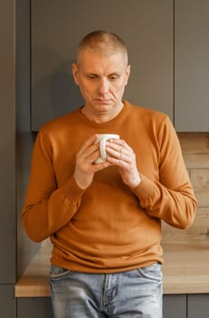 Portrait of a mature man in an orange jumper in the kitchen with a mug of warm tea. Vertical frame.