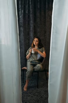 A young woman sits in a chair with a mug of tea and a tablet in her hands. Vertical frame.
