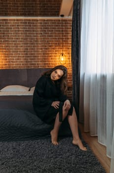 Portrait of a beautiful woman in a black bathrobe, who is sitting on the bed, looking at the camera.