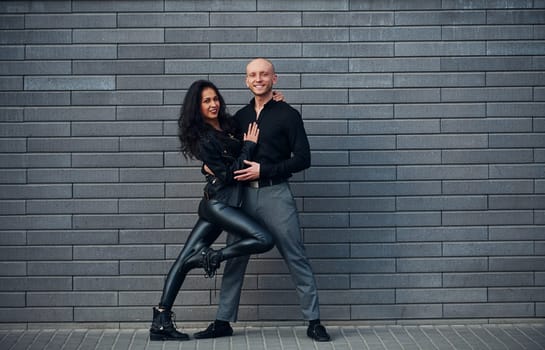 Beautiful couple in black clothes dancing together against wall outdoors.