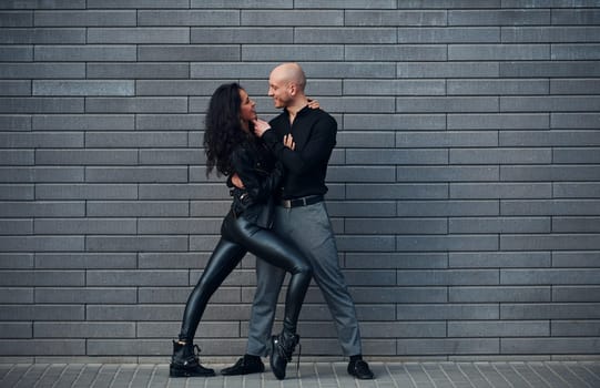 Beautiful couple in black clothes dancing together against wall outdoors.