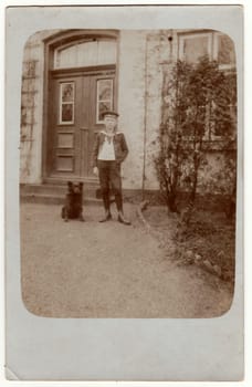 HAMBURG, GERMANY - DEUTSCHES REICH - CIRCA 1920s: Vintage photo shows boy poses in front of house. Boy wears a sailor costume and stands next to dog. Retro black and white photography. Circa 1920s.