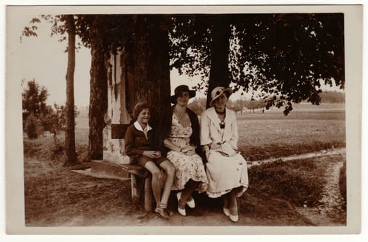 THE CZECHOSLOVAK REPUBLIC - CIRCA 1930s: Vintage photo shows women wear elegant dresses and women's parisisol big brim hat. The women and boy pose outdoors and sit on the bench. Retro black and white photography. Circa 1930s