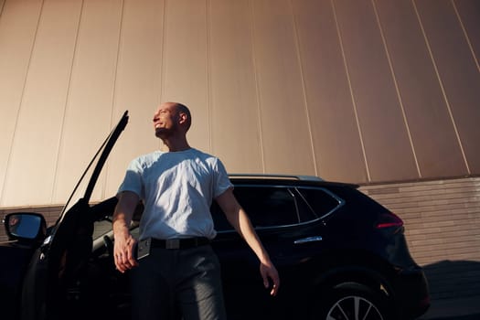 Bald man standing near his modern black car outdoors at daytime.