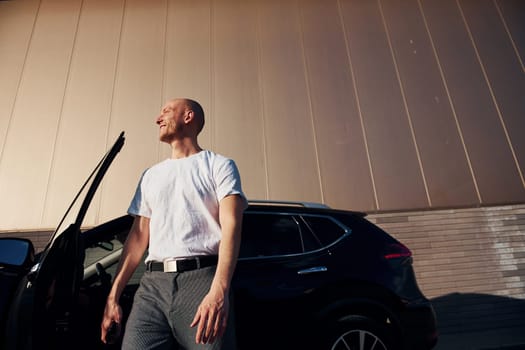 Bald man standing near his modern black car outdoors at daytime.