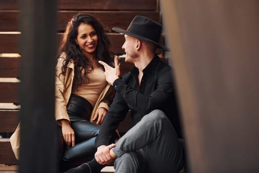 Cheerful brunette in black leggings sitting on wooden stairs with her man in hat.