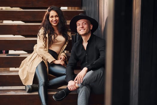 Cheerful brunette in black leggings sitting on wooden stairs with her man in hat.