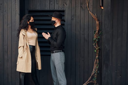 Woman with black curly hair standing against black wooden building exterior with her man in hat.