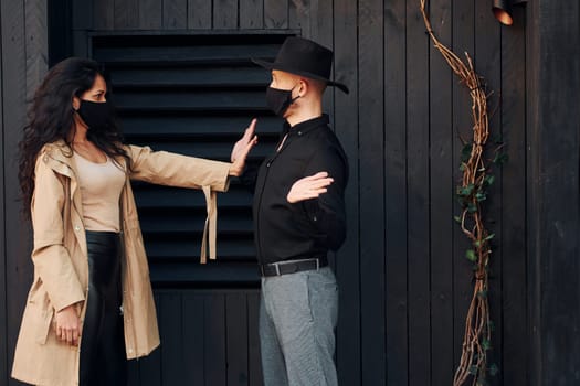 Woman with black curly hair standing against black wooden building exterior with her man in hat.