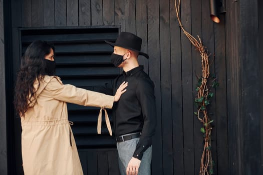 Woman with black curly hair standing against black wooden building exterior with her man in hat.