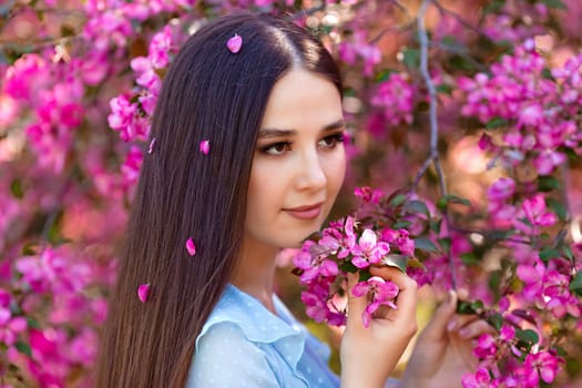 A pretty brunette girl with pink petals in long hair is standing near a pink blooming apple tree, looking away. Close up.