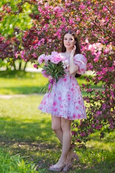 Adorable girl, stands with a large bouquet of peonies, near pink flowering trees in the garden, in spring on a sunny day. Copy space. Vertical