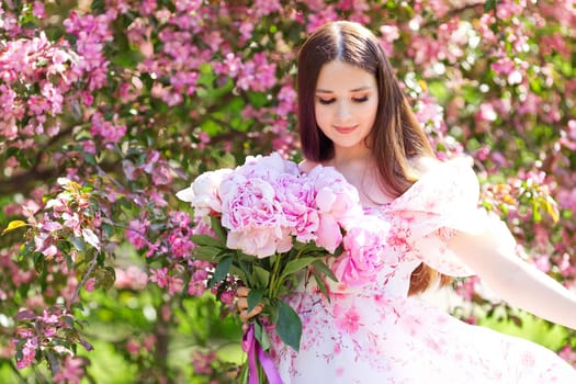 A girl in a light pink dress dancing with a large bouquet of pink peonies in a pink blooming garden on a sunny day. close up. Copy space