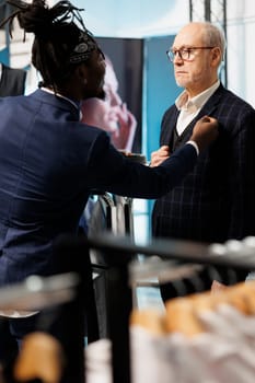 African american employee helping stylish man with suit in clothing store, client looking to buy modern clothes. Senior customer buying fashionable merchandise and trendy accessories in showroom
