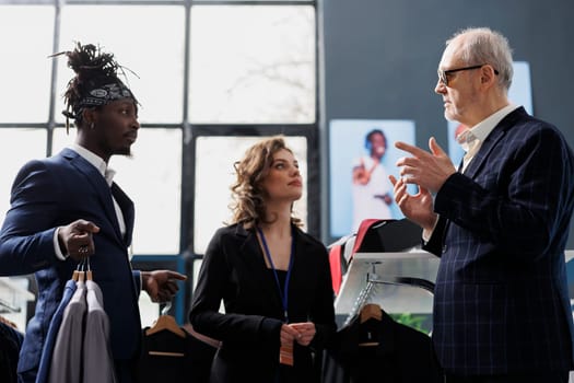 Diverse workers helping senior client with trendy shirt, choosing stylish clothes for casual wear in clothing store. Elderly man buying fashionable merchandise and accessories in modern boutique