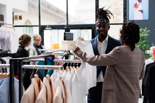 Showroom employee helping african american client with choosing white shirt during commerical activity. Elegant customer buying fashionable clothes in shopping centre. Fashion concept