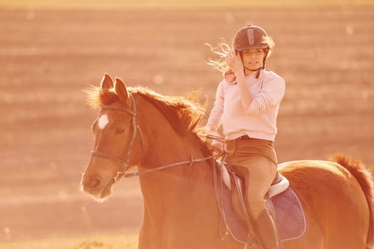 Young woman in protective hat riding her horse in agriculture field at sunny daytime.