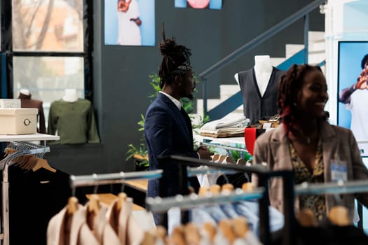 Elegant man looking at trendy clothes on hangers in showroom, shopping for fashionable merchandise to increase wardrobe. African american customer buying new fashion collection in modern boutique