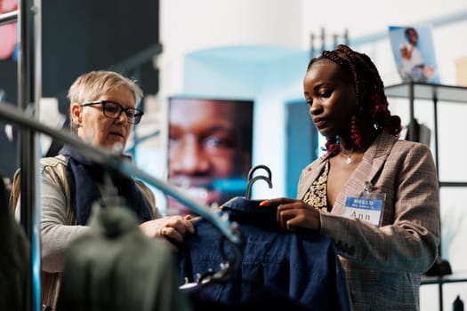 African american worker helping senior customer choosing shirt for husband in modern boutique. Elderly woman shopping for casual wear, buying fashionable merchandise in clothing store