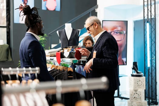 African american worker helping customer with casual wear, discussing tie fabric in clothing store. Elderly man shopping for formal suit, buying fashionable clothes and stylish merchandise