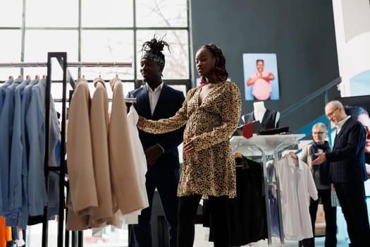 Pregnant woman looking at shirt checking material in showroom, african american couple shopping for pregnancy clothes. Clients buying maternity merchandise in clothing store. Fashion concept