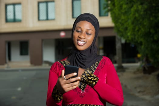 young muslim woman standing outdoors looking at cell phone while smiling.