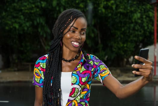 young african girl under a light rain watching mobile phone while laughing.