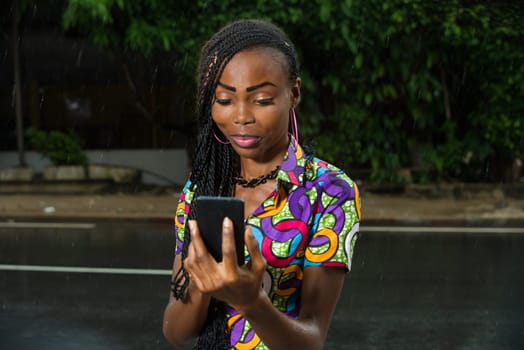 young girl under a light rain looking at mobile phone while smiling.