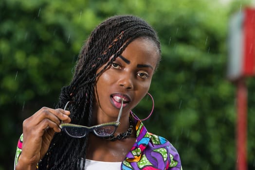 young girl under a light rain watching the camera with glasses in her mouth.