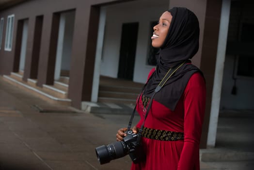 young muslim woman standing outdoors with camera looking in front of her smiling.