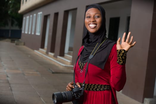 young muslim woman standing outdoors with camera greeting with laughter.
