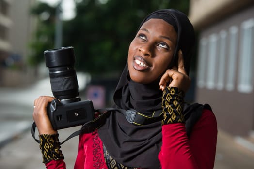 young muslim woman standing outdoors with a camera thinking about looking up there.
