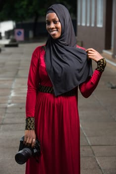 young muslim woman standing outdoors with camera smiling looking at the camera.