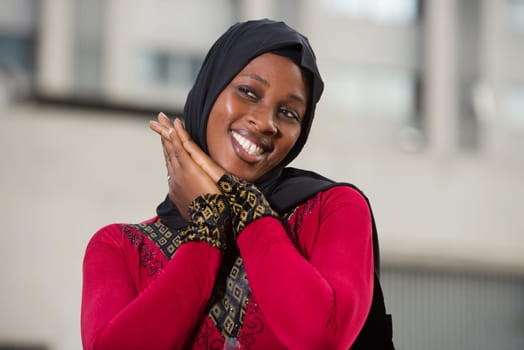 young muslim woman standing outdoors looking at the camera laughing with her hands clasped together.