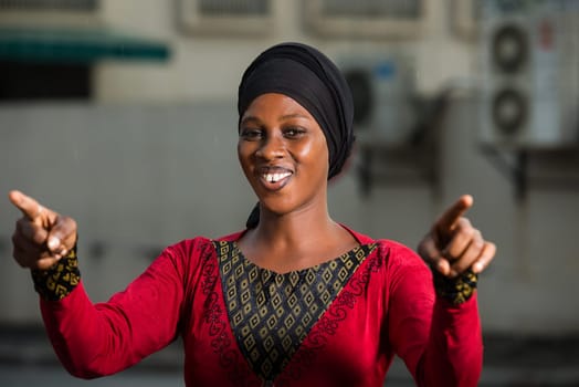 young woman standing in headscarf outdoors smiling to make finger gesture.