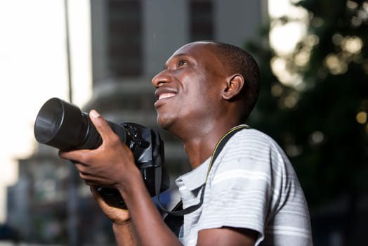 young man standing outdoors looking up with camera in hand smiling.