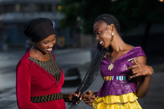 young african woman standing outdoors contemplating the braids of her girlfriend smiling.