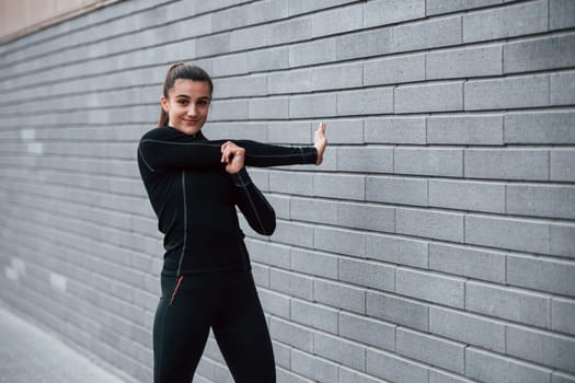 Young sportive girl in black sportswear outdoors doing stretching near gray wall.