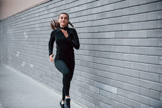 Young sportive girl in black sportswear running outdoors near gray wall.