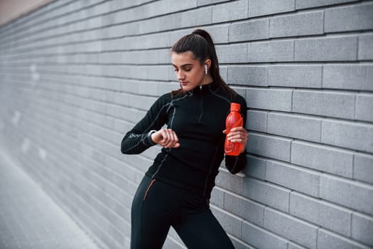 Young sportive girl in black sportswear standing outdoors near gray wall with bottle of water and taking a break.