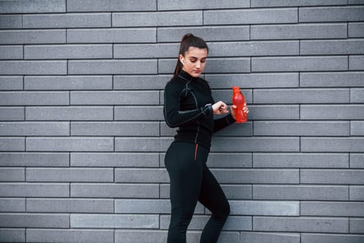 Young sportive girl in black sportswear standing outdoors near gray wall with bottle of water and taking a break.