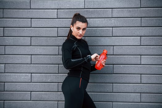 Young sportive girl in black sportswear standing outdoors near gray wall with bottle of water and taking a break.