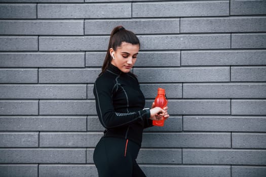 Young sportive girl in black sportswear standing outdoors near gray wall with bottle of water and taking a break.