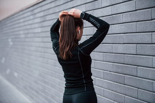 Young sportive girl in black sportswear standing outdoors near gray wall.