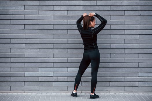 Rear view of young sportive girl in black sportswear that standing outdoors near gray wall.