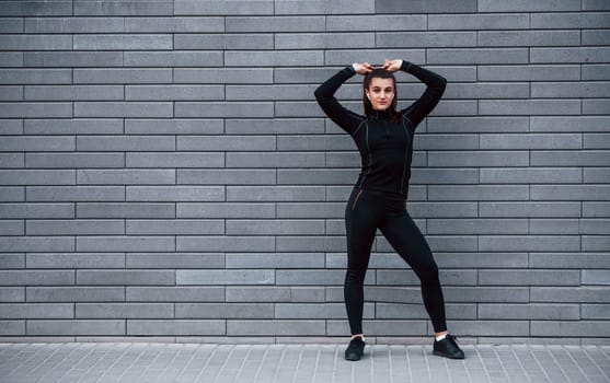 Young sportive girl in black sportswear standing outdoors near gray wall.