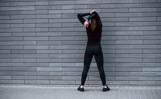 Rear view of young sportive girl in black sportswear that standing outdoors near gray wall.