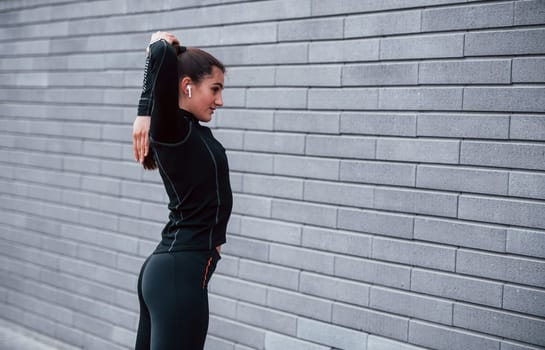 Young sportive girl in black sportswear standing outdoors near gray wall.