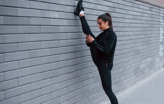 Young sportive girl with phone in black sportswear doing legs stretching outdoors by using gray wall.